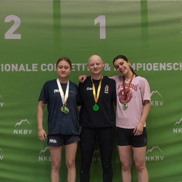 Drie jonge vrouwen staan op een podium voor een groene achtergrond, met medailles om. De vrouw in het midden is de winnares van de eerste plaats, geflankeerd door de nummers twee en drie; ze glimlachen allemaal naar de camera.